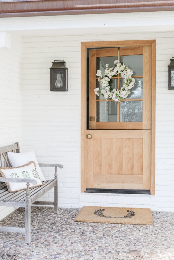 A wooden front door with glass panels and a floral wreath, next to a bench with a cushion and outdoor pillows on a house with white brick walls.