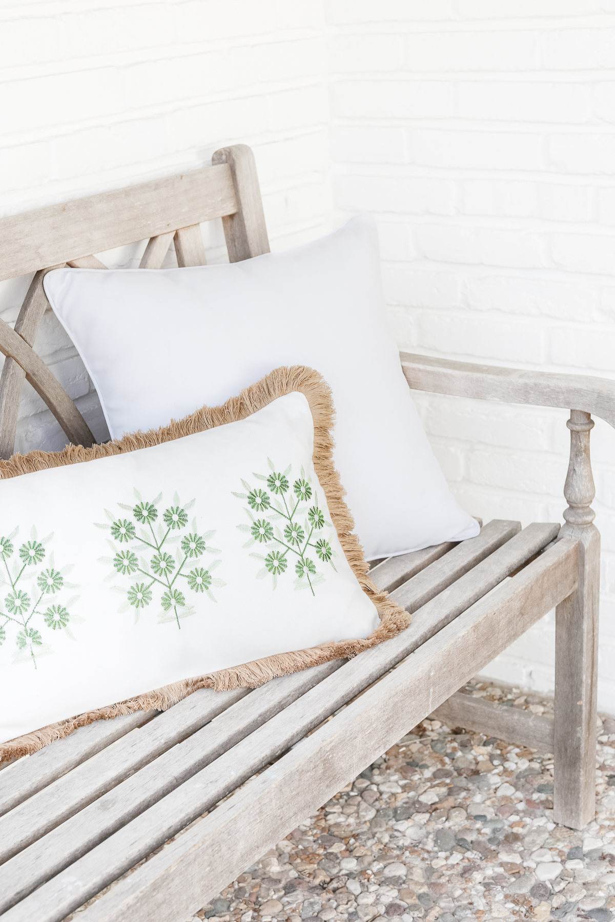 A wooden bench with a fringed white outdoor pillow and another outdoor pillow with green botanical print against a white brick wall.