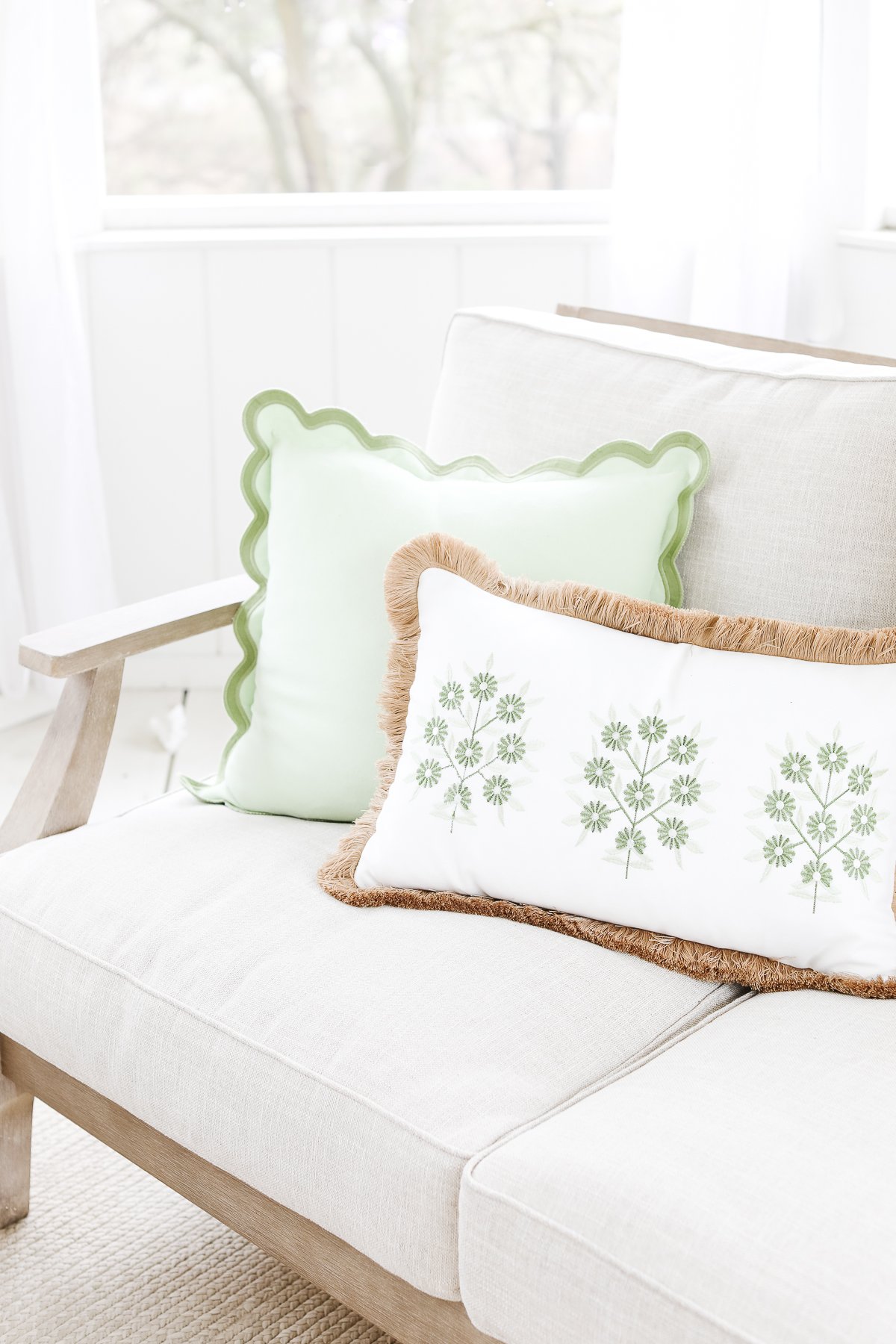 A bright and cozy corner with a beige cushioned bench, decorated with outdoor pillows: one green scalloped-edge pillow and one white pillow with green botanical print.