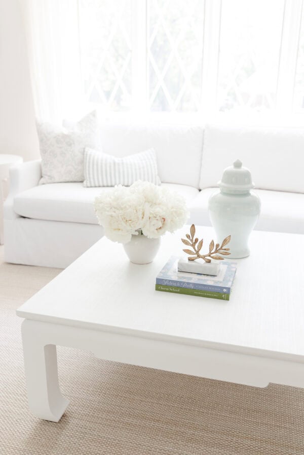 A white coffee table with a vase of white flowers, two stacked books, a decorative gold leaf, and a pale blue jar sits atop a custom sisal rug with border in front of a white sofa in a bright living room.