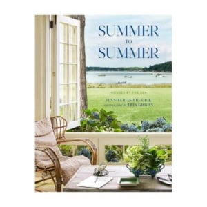 Book cover for "Summer to Summer: Houses by the Sea" featuring an open window view of a coastal landscape, a wicker chair, and a table adorned with decorative books and plants.