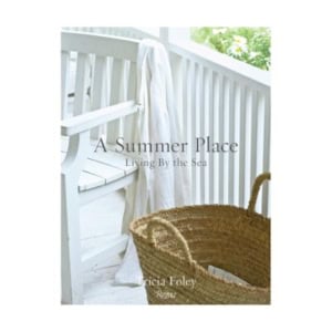 A white bench with a draped cloth beside a wicker basket on a porch, featuring decorative books including the title "A Summer Place: Living By the Sea" by Tricia Foley overlaid.