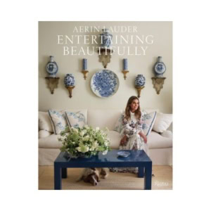 A woman sits on a white sofa in a tastefully decorated living room with blue accents, floral arrangements, decorative books, and plates and vases adorning the wall.