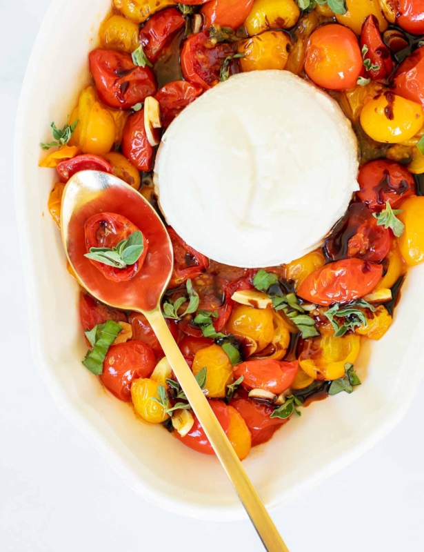 Overhead view of blistered tomatoes on serving platter with burrata, with single tomato and basil leaves on spoon