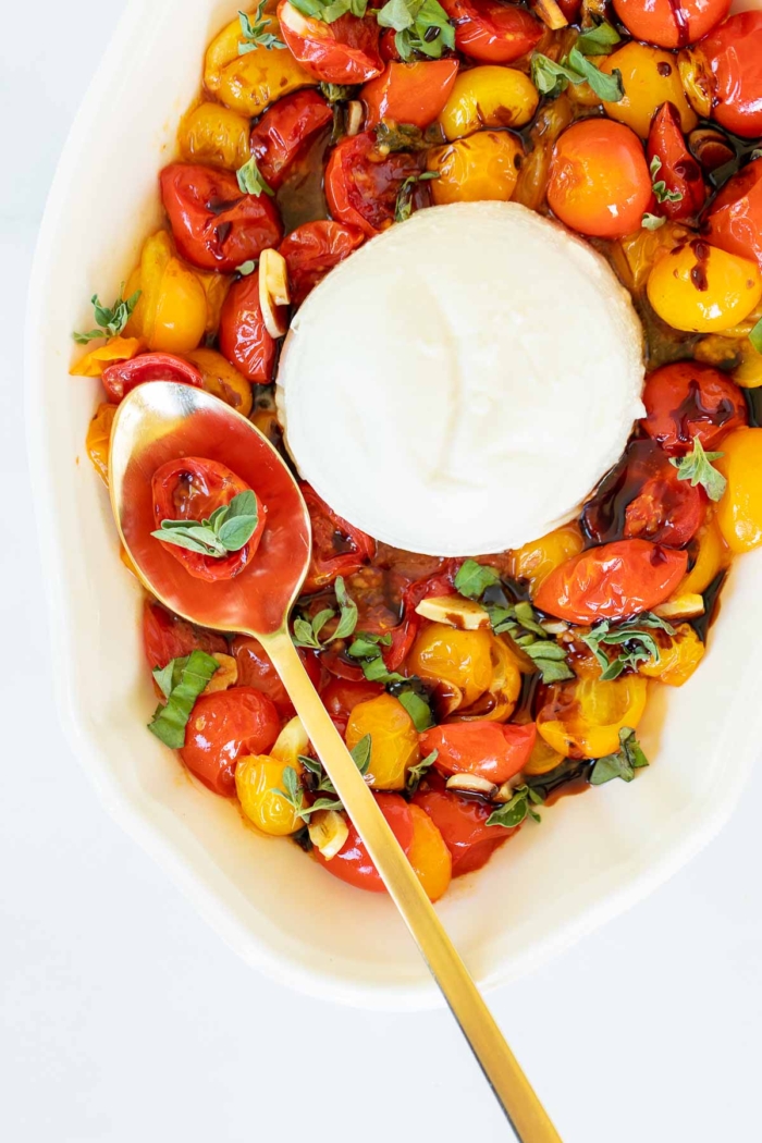 Overhead view of blistered tomatoes on serving platter with burrata, with single tomato and basil leaves on spoon