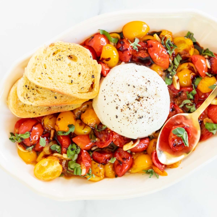 Overhead view of blistered tomatoes on platter with 3 crostini and ball of burrata