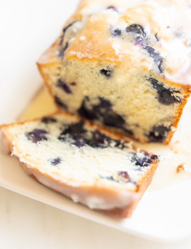 A loaf of blueberry bread, topped with a glaze. One slice is cut at the end, and it's on a white platter.
