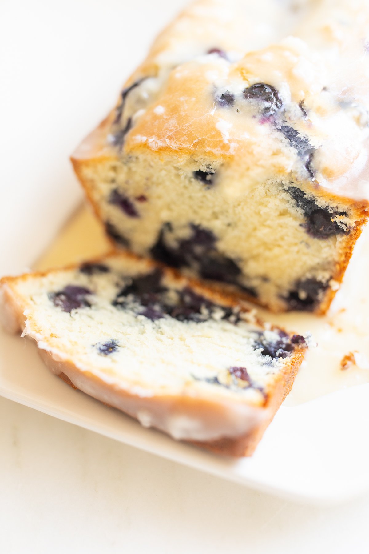 A loaf of blueberry bread, topped with a glaze. One slice is cut at the end, and it's on a white platter.