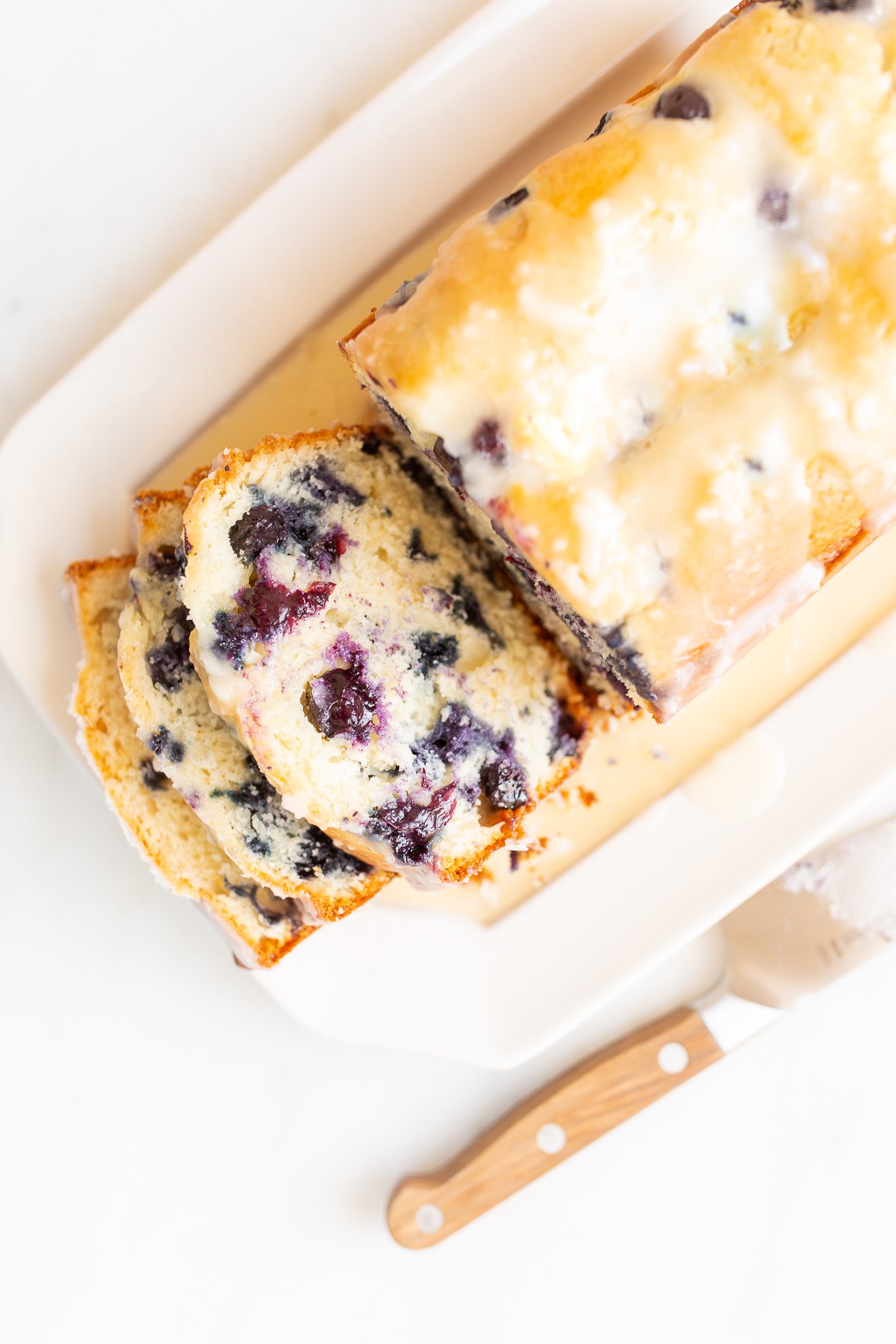 A loaf of blueberry bread, topped with a glaze. Two slices are cut at the end, and it's on a white platter.