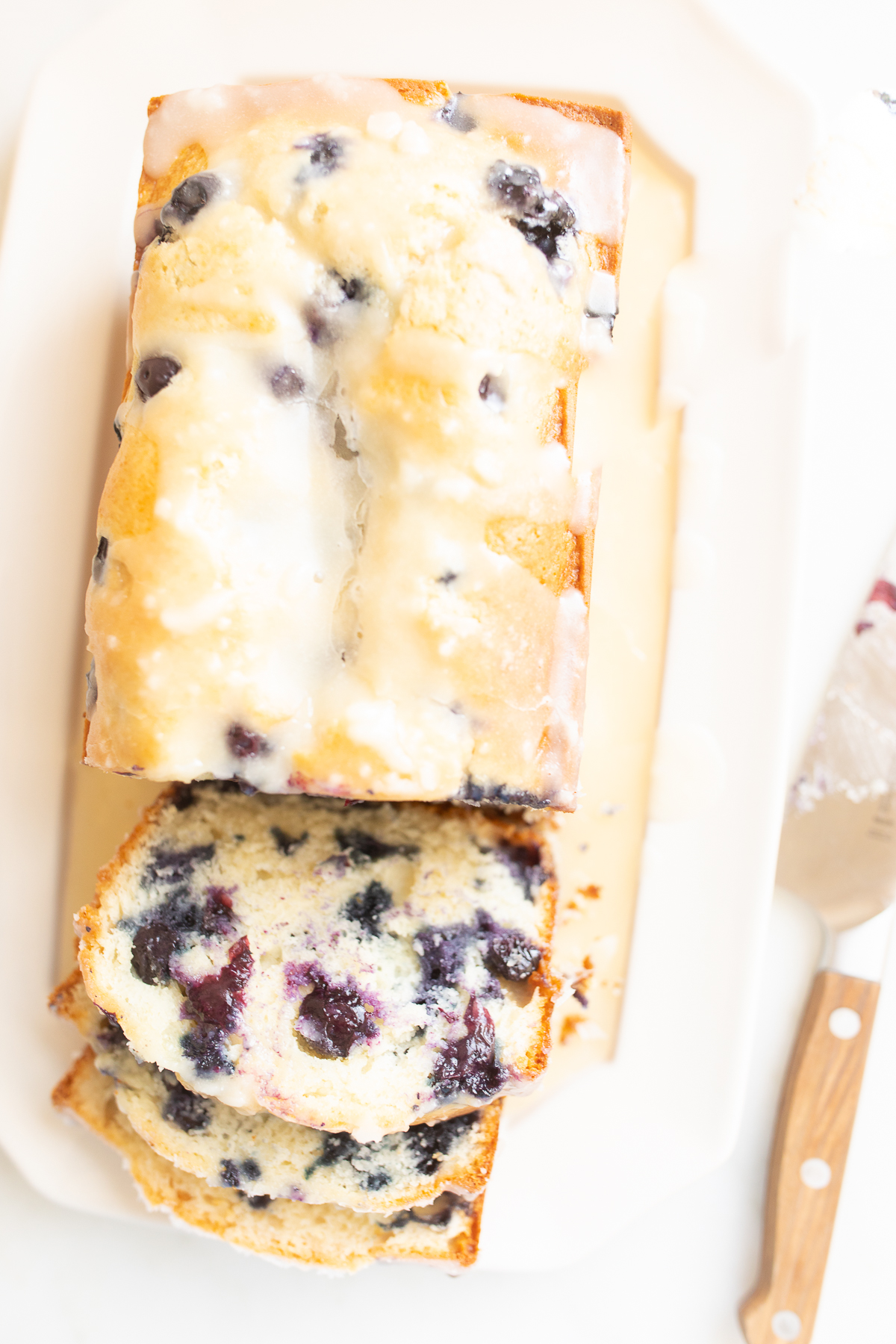 A loaf of blueberry bread, topped with a glaze. Two slices are cut at the end, and it's on a white platter.