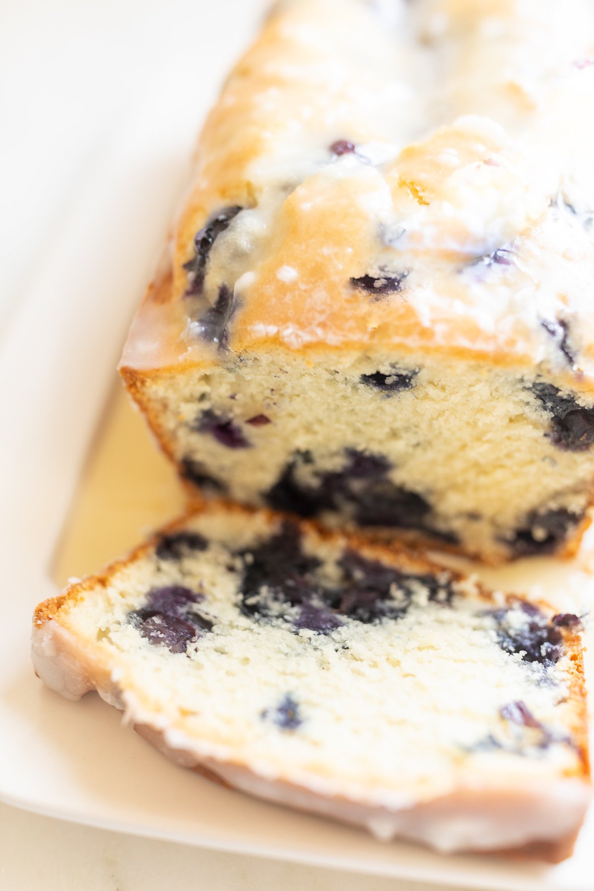 A loaf of blueberry bread, topped with a glaze. One slice is cut at the end, and it's on a white platter. 