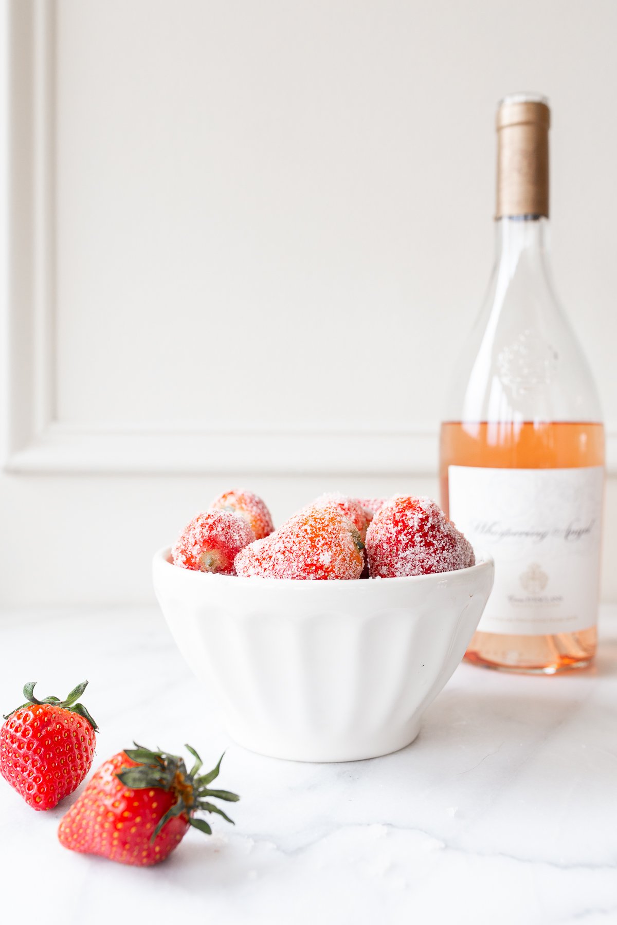 Drunken strawberries rolled in sugar in a white bowl, with a bottle of wine in the background. 