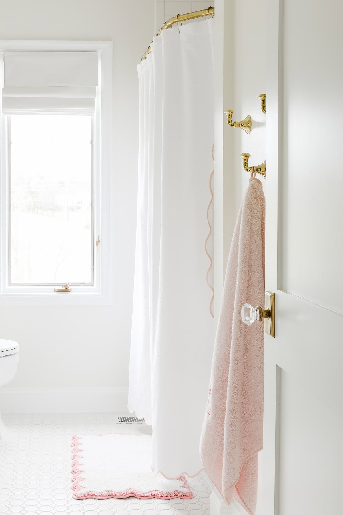 A bright bathroom interior with a white shower curtain, a hanging pink towel, and a white door partially open, revealing brass door knobs, a toilet and a window with a view outside.