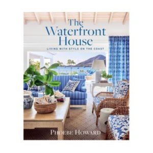 Book cover of "The Waterfront House" by Phoebe Howard, featuring a coastal-themed living room with blue and white decor, decorative books on the coffee table, and large windows overlooking the water.