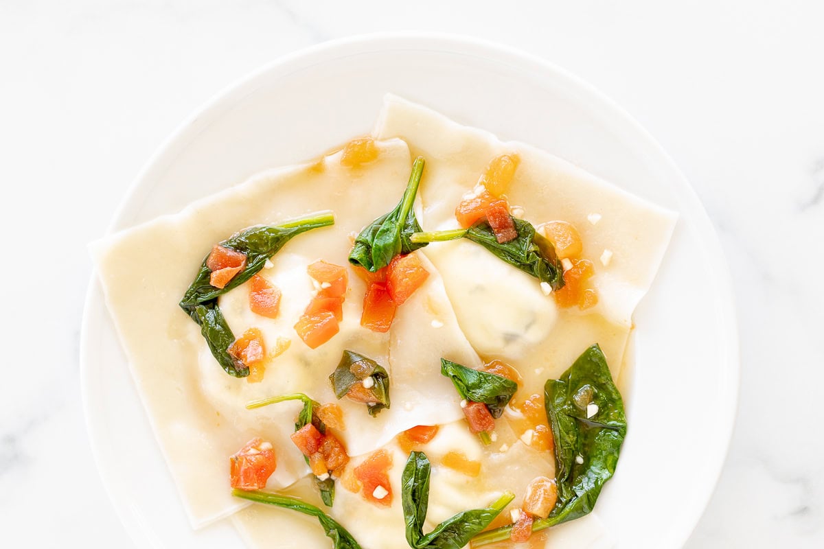 A plate of goat cheese ravioli topped with chopped tomatoes and spinach leaves, served on a white marble background.