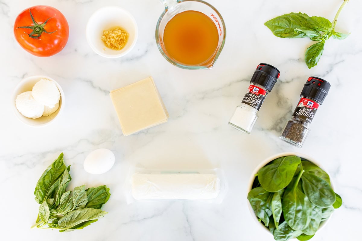 Overhead view of a white marble countertop with ingredients for cooking: tomato, basil, mozzarella, spices, olive oil, goat cheese ravioli, and an egg, neatly arranged.