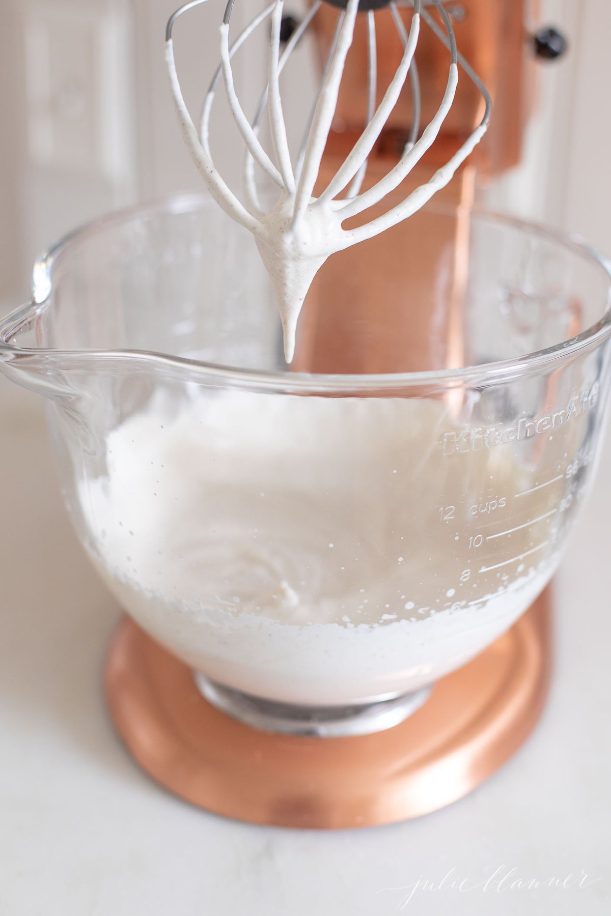 A clear glass mixing bowl of a stand mixer filled with whipped cream.