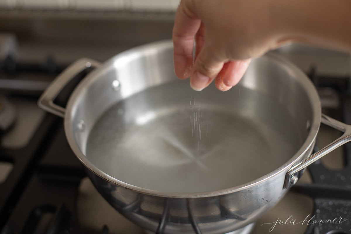 A hand adding salt to a stainless steel pot of water in a guide for how to cook pasta. 