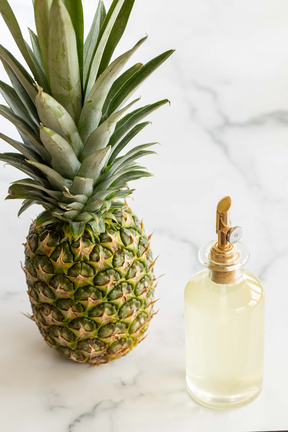 A fresh pineapple on a marble countertop next to a bottle of simple syrup.