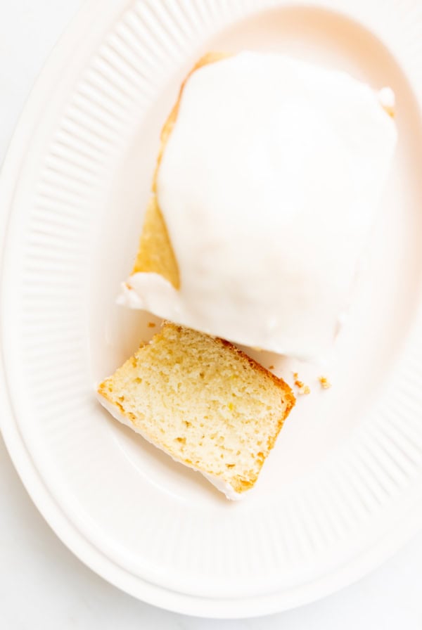 A slice of lemon bread with white icing on a ribbed white plate, with one corner of the cake already cut and placed beside it.