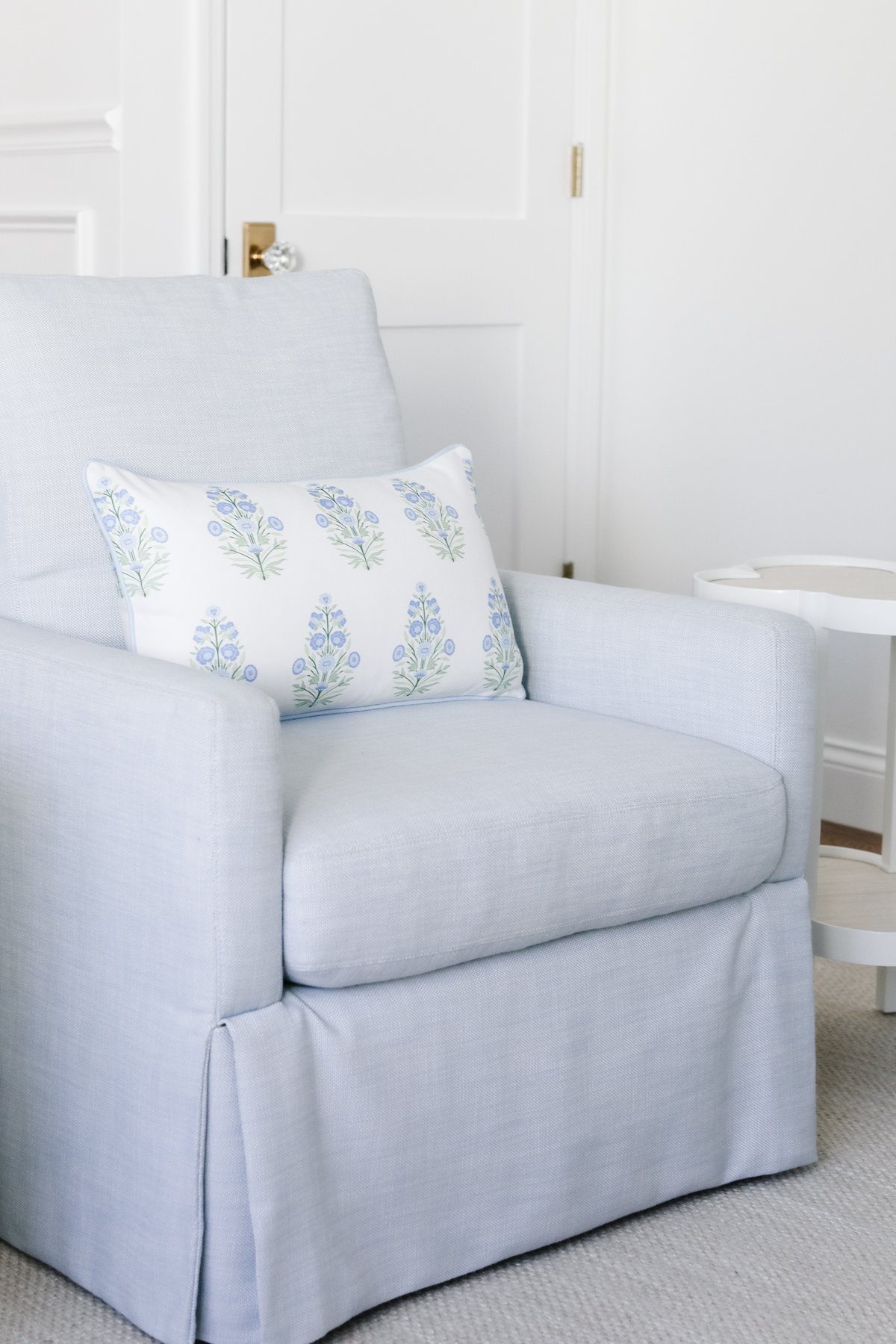 A light blue armchair with a floral-patterned pillow, enhanced by plush pillow inserts, sits elegantly in a room with a white door and a small white side table.