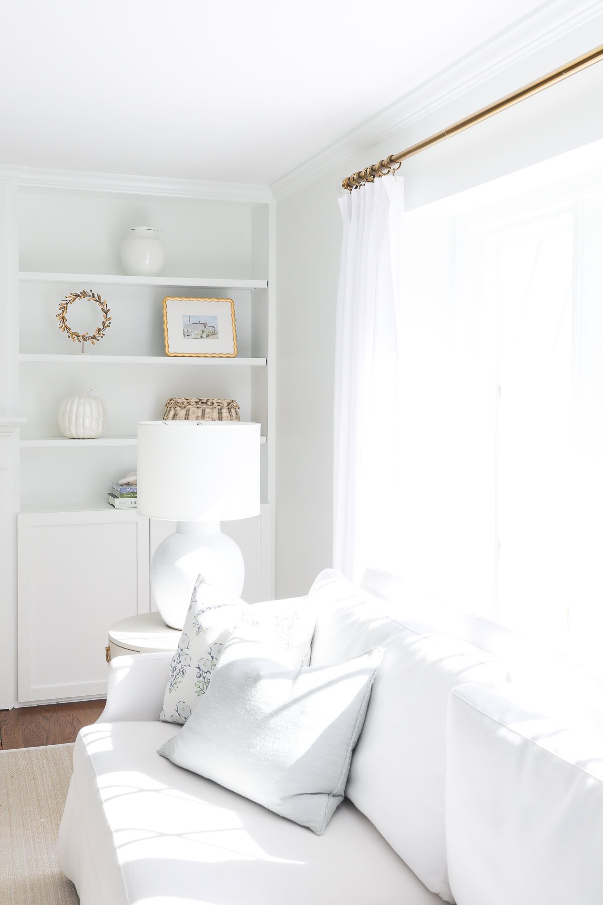 Bright living room with a white sofa, light blue pillow inserts adding a pop of color, a lamp, and a shelf with decor, including a framed picture and a vase, against a crisp white wall.