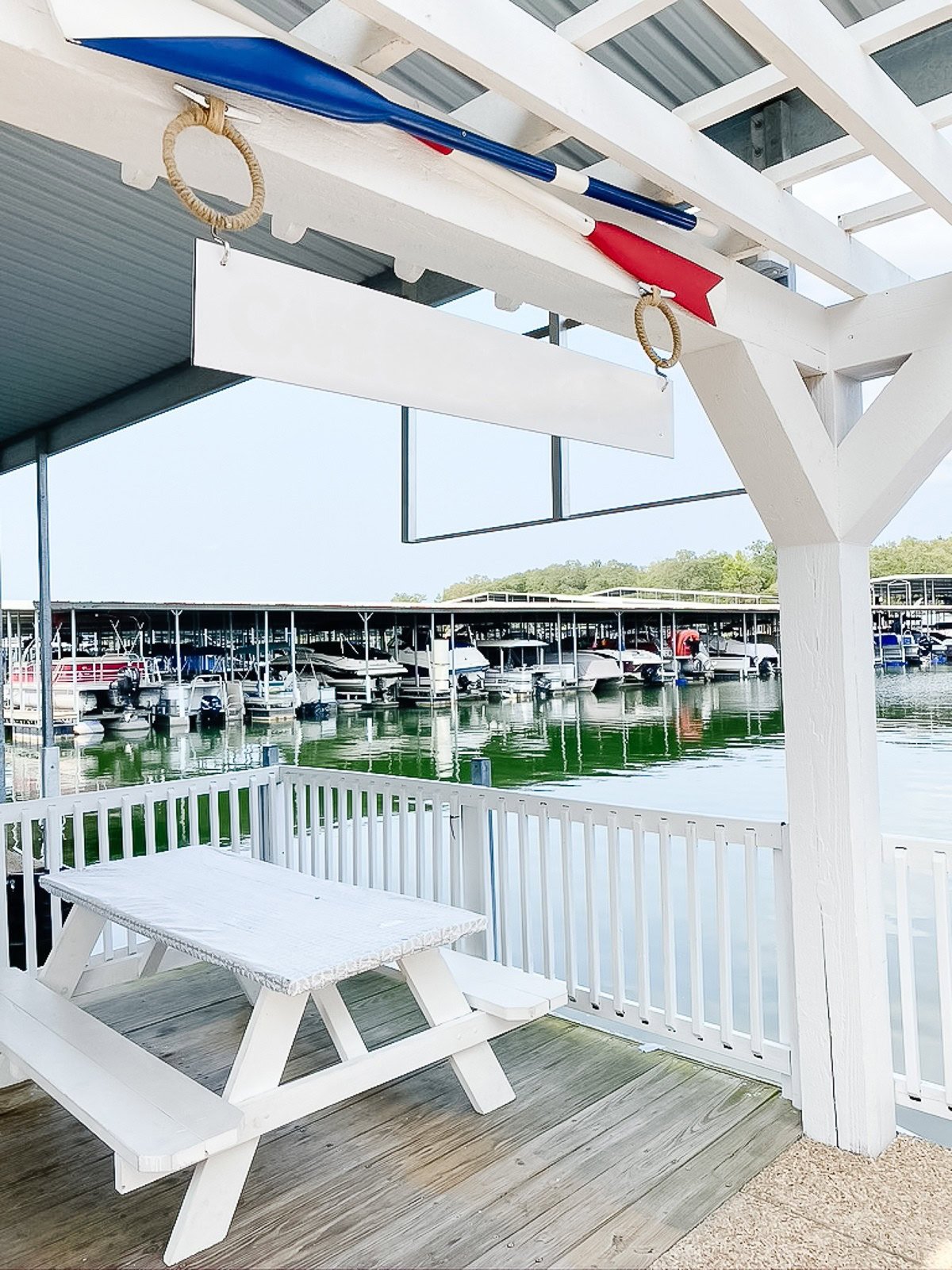 A white picnic table sits on a wooden dock under a shaded pergola, overlooking a marina with docked boats and calm green water—evoking the serene ambiance found at some Table Rock Lake restaurants.
