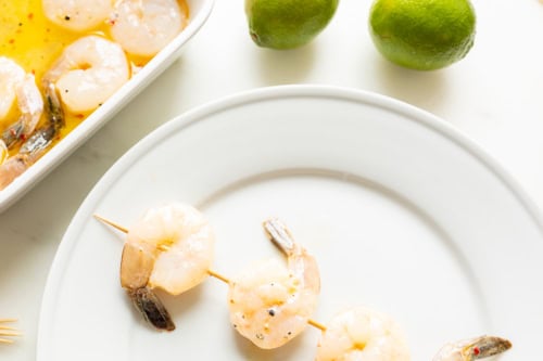 Tequila lime shrimp skewers on a white plate next to a baking dish with more shrimp and two limes.