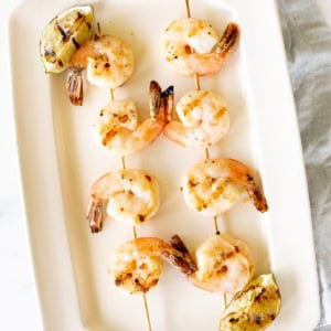 Two skewers of tequila lime shrimp with charred lemon wedges, displayed on a white rectangular plate.