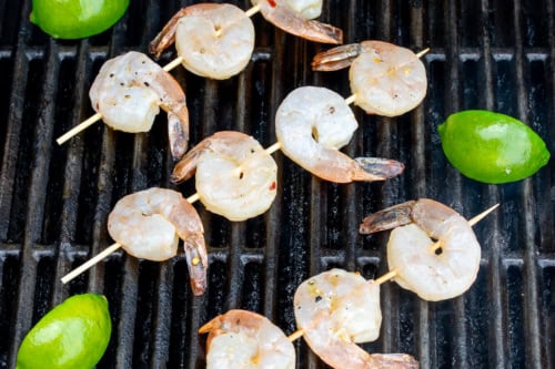 Shrimp skewers and halved limes sizzle on the grill, with the tequila lime shrimp turning slightly opaque as they cook.
