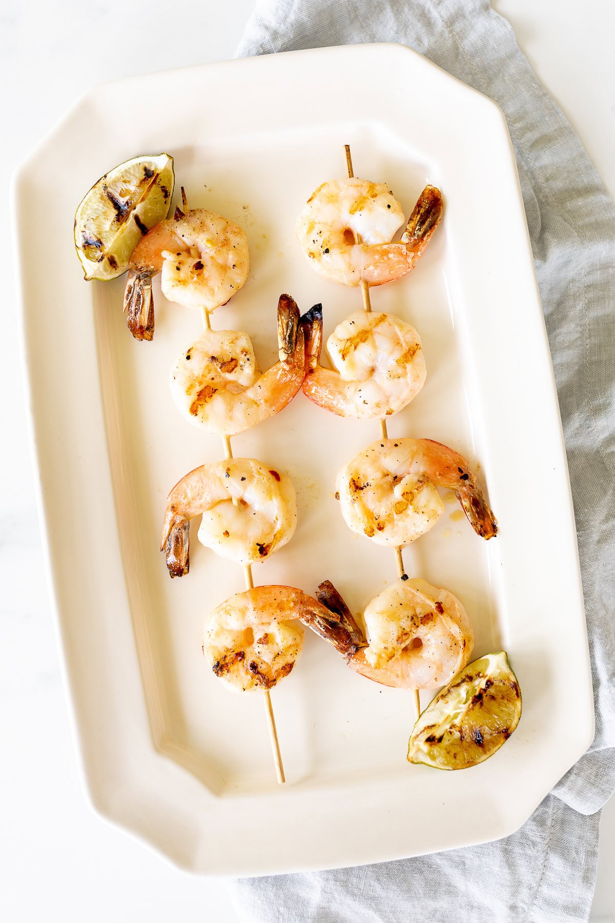 Grilled tequila lime shrimp skewers with lemon wedges on a rectangular white plate.