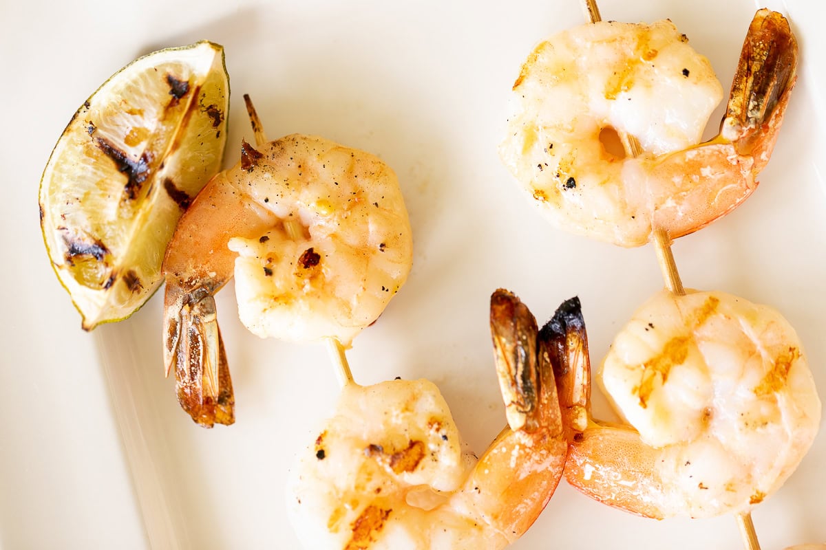 Close-up of tequila lime shrimp skewers with a wedge of charred lime on a white plate.