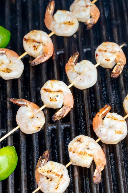 Tequila lime shrimp skewers grilling on a barbecue with lime wedges on the side.
