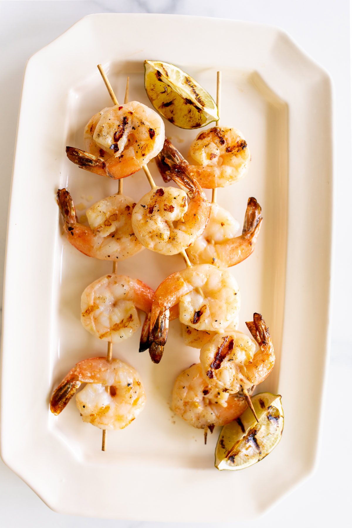Two skewers of tequila lime shrimp garnished with grilled lemon wedges are served on a white rectangular plate.