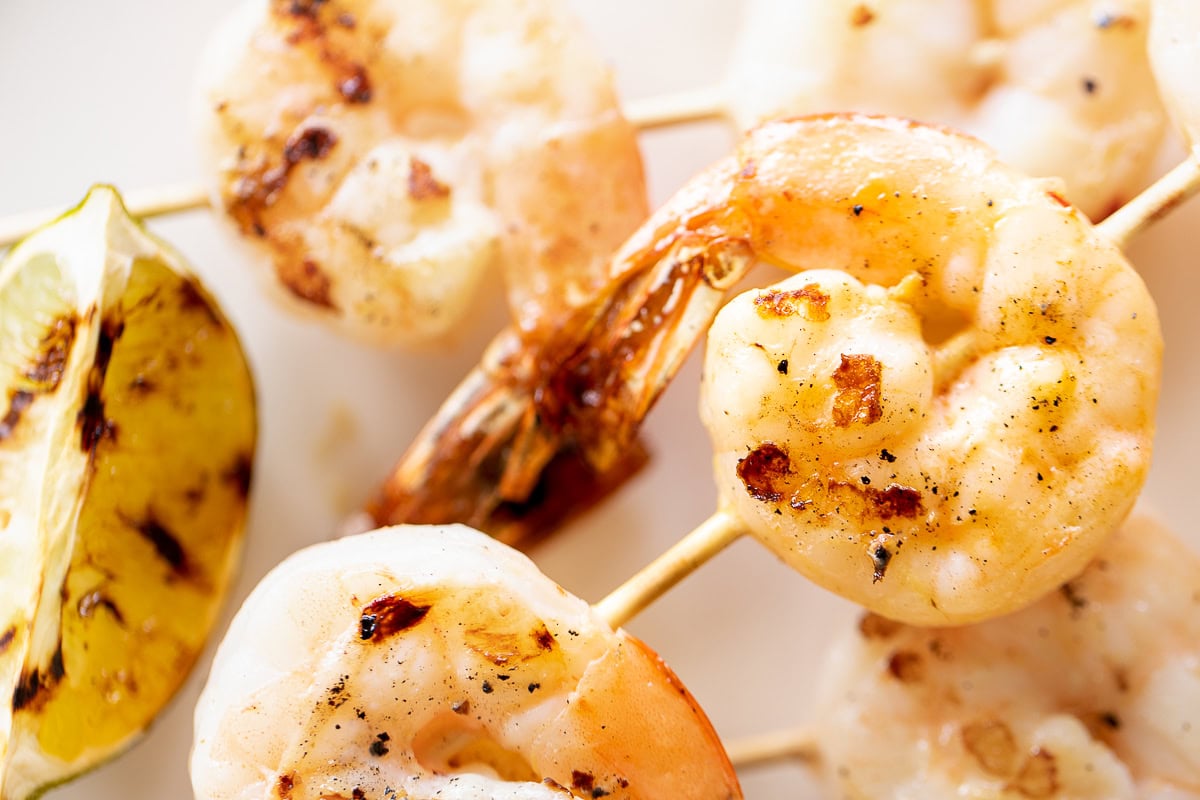 Close-up of tequila lime shrimp skewers and a charred lemon wedge on a white plate. The shrimp are lightly browned with visible grill marks.