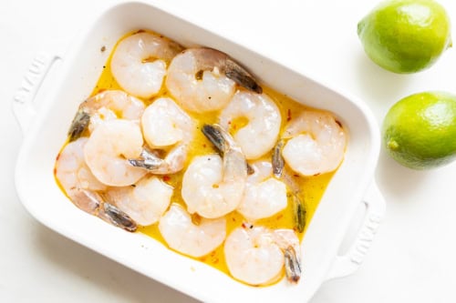A white dish filled with raw tequila lime shrimp marinating in a yellowish liquid next to two whole limes.