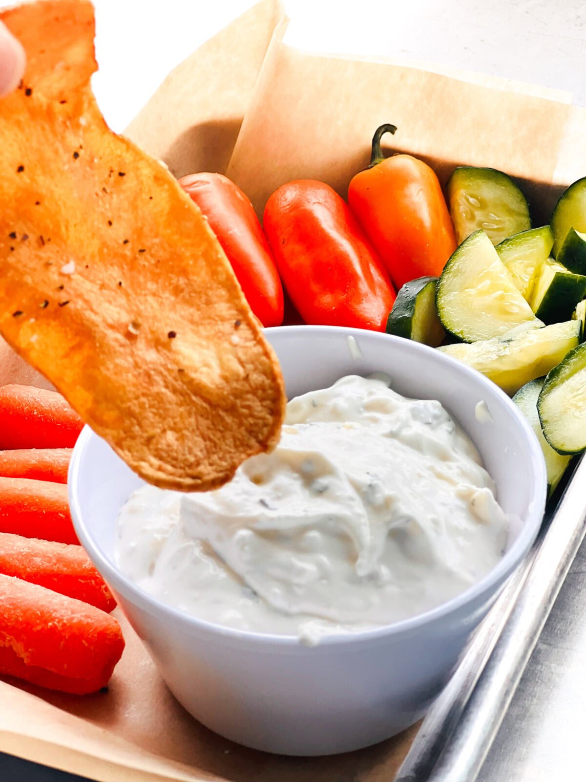 A hand holds a crisp chip above a bowl of white dip, with sliced carrots, tomatoes, and cucumbers arranged beside it on a tray—perfect for snacking at one of the scenic Table Rock Lake restaurants.