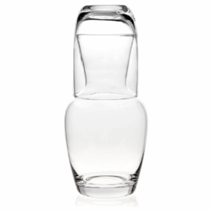 A clear glass cup is placed upside down on a matching clear glass carafe, both empty, creating an inviting touch perfect for a guest room, against a white background.