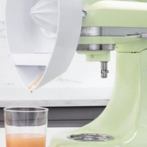 A green kitchen gadget stand mixer with an attached pouring shield dispensing liquid into a glass.