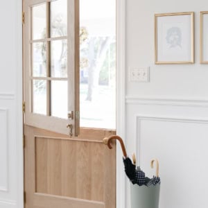A wooden Dutch door, painted in Sherwin Williams Extra White, is half open in a bright entryway. An umbrella stand with umbrellas sits nearby, and framed art hangs on the crisp white wall.
