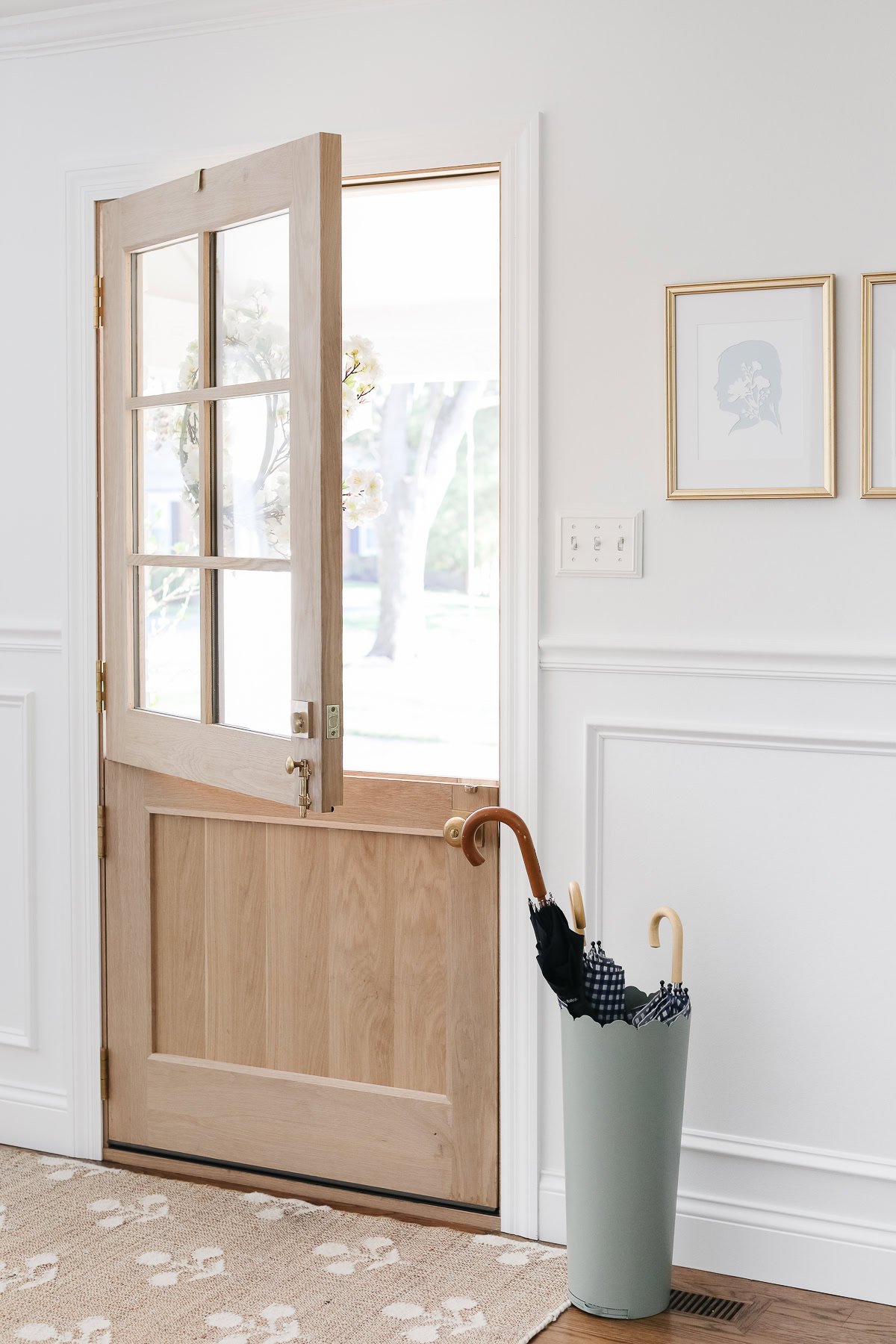 A wooden Dutch door, painted in Sherwin Williams Extra White, is half open in a bright entryway. An umbrella stand with umbrellas sits nearby, and framed art hangs on the crisp white wall.