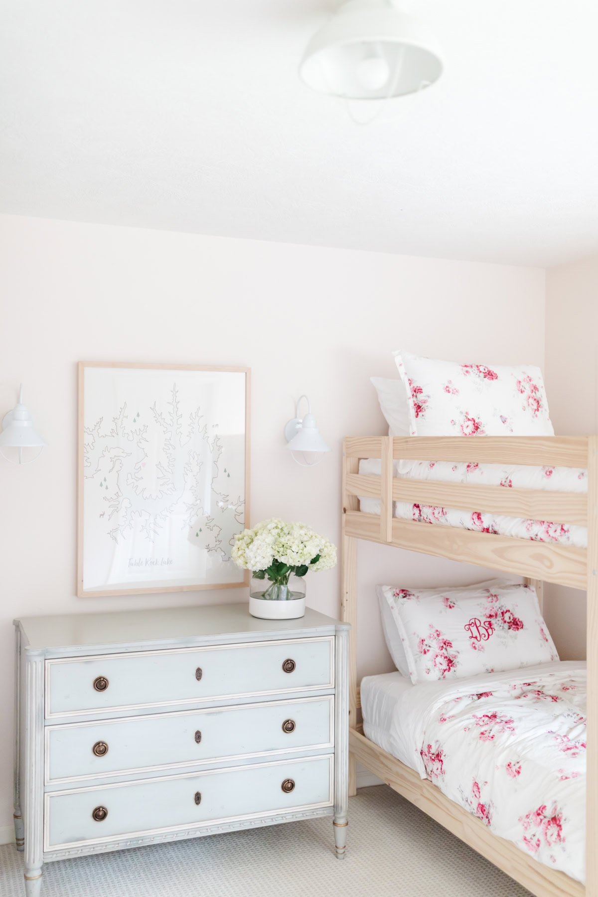 A light-filled bedroom with blush pink paint, a wooden bunk bed, floral bedding, a white dresser with brass knobs, a vase of white flowers, and framed wall art.