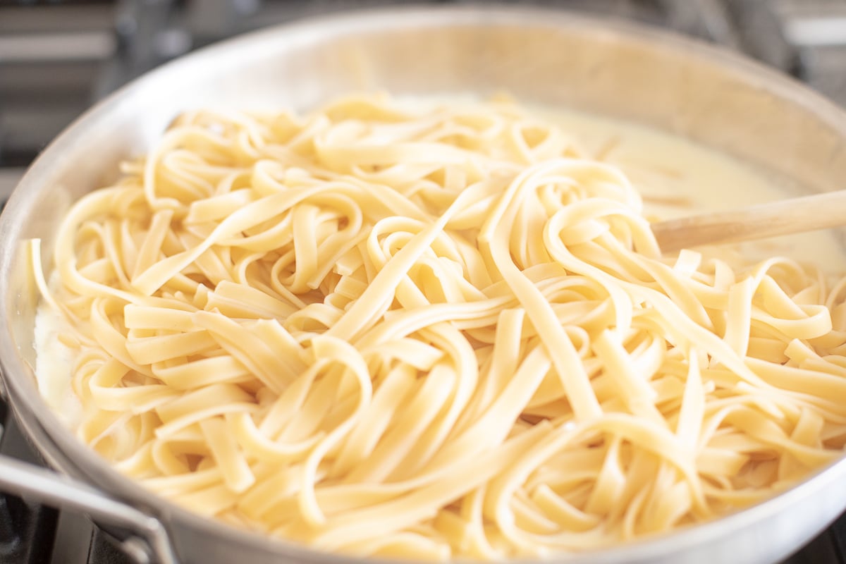 A stainless steel pan on a stovetop, filled with pasta. Image is part of a tutorial on how to reheat pasta.