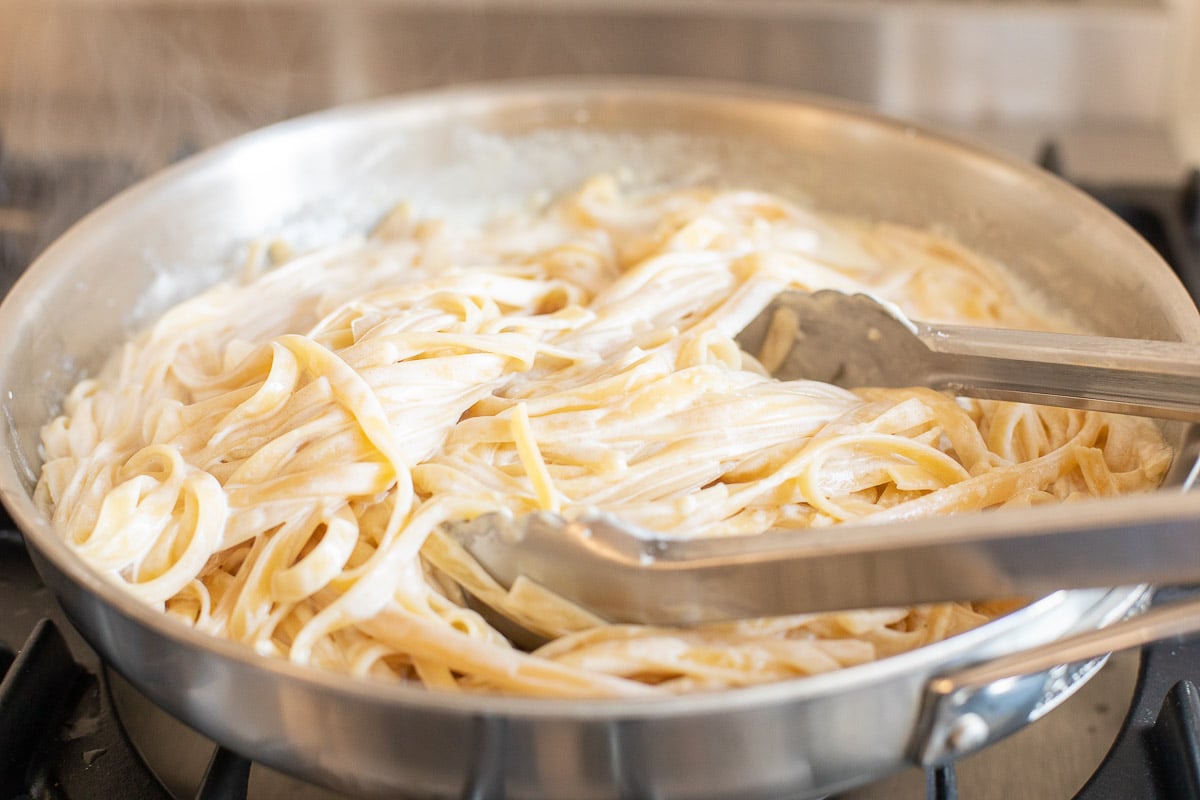 A stainless steel pan on a stovetop, filled with pasta. Image is part of a tutorial on how to reheat pasta.