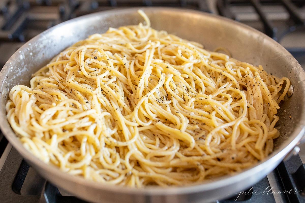 A stainless steel pan on a stovetop, filled with pasta. Image is part of a tutorial on how to reheat pasta.