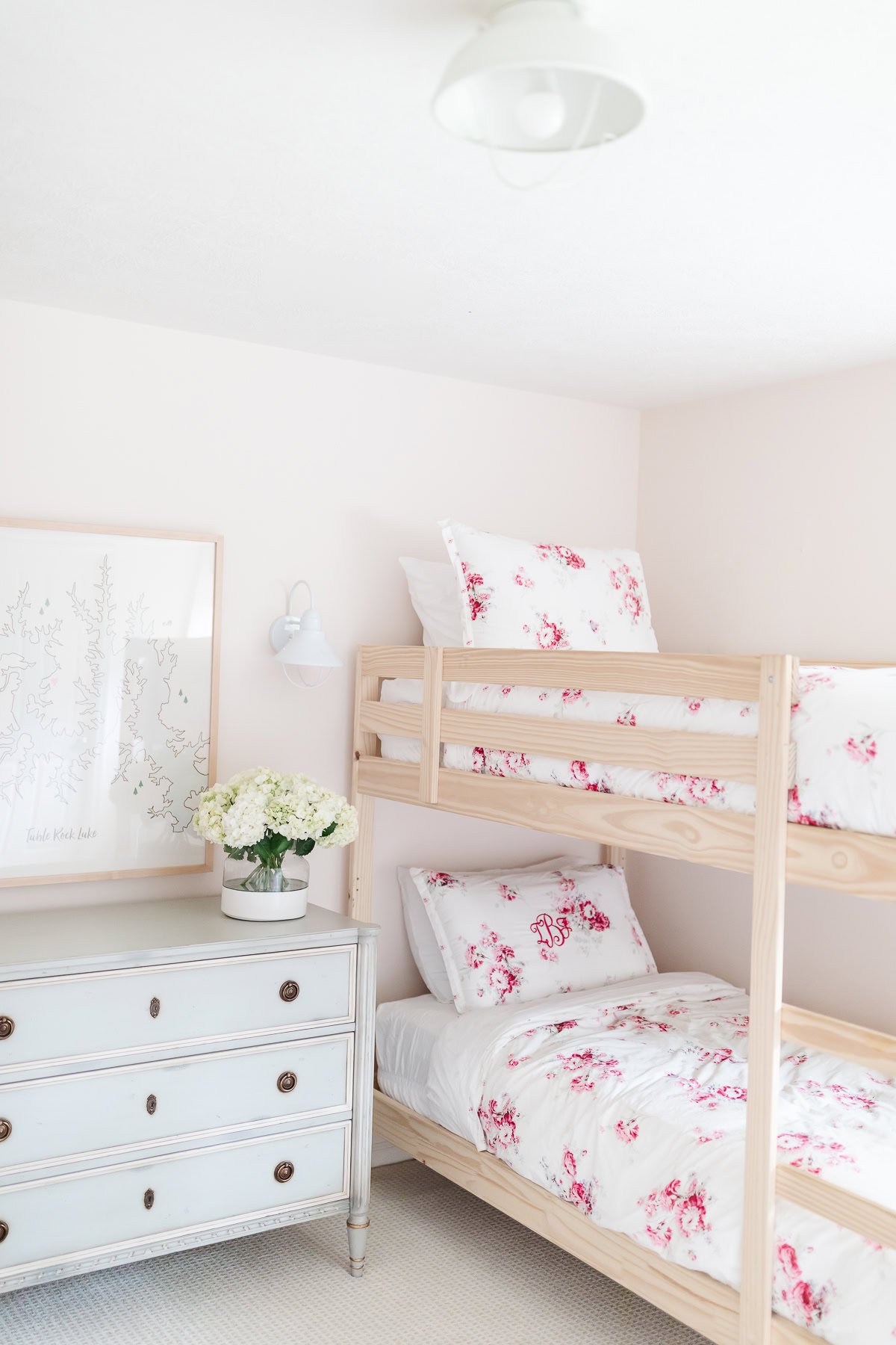 A light-filled bedroom with wooden bunk beds dressed in white and pink floral bedding, blush pink paint on the walls, a vintage white dresser, a vase of white flowers, and a framed wall art.