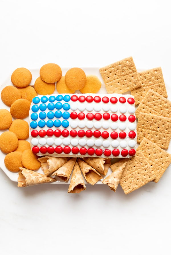 A rectangular dessert decorated to resemble the U.S. flag with red, white, and blue candy, surrounded by vanilla wafers, graham crackers, and ice cream cones on a white surface. This is one of those quintessential red white and blue desserts perfect for patriotic celebrations.