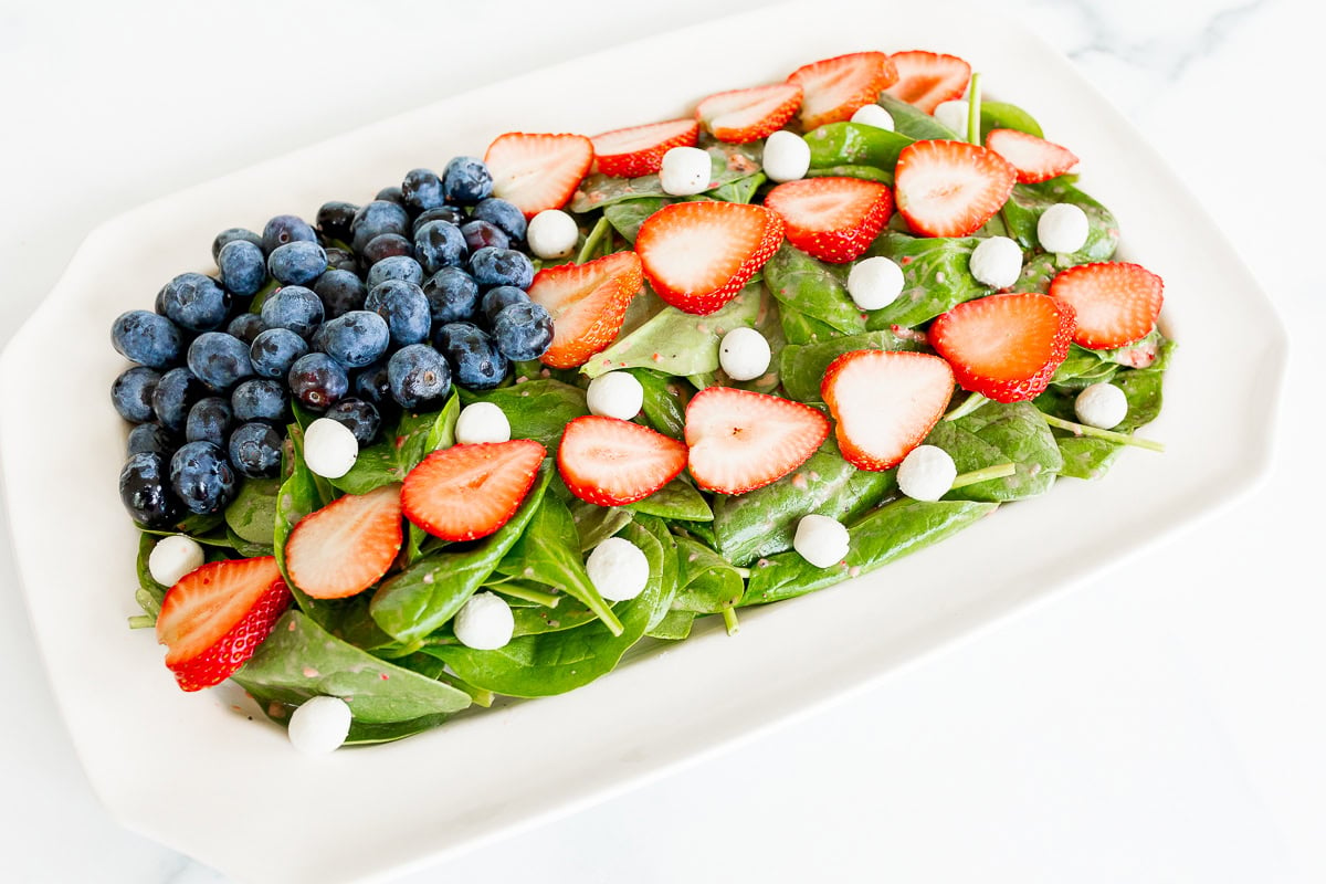 A vibrant red, white, and blue salad with fresh spinach, sliced strawberries, whole blueberries, and white round candies, all arranged in a white rectangular dish on a marble surface.