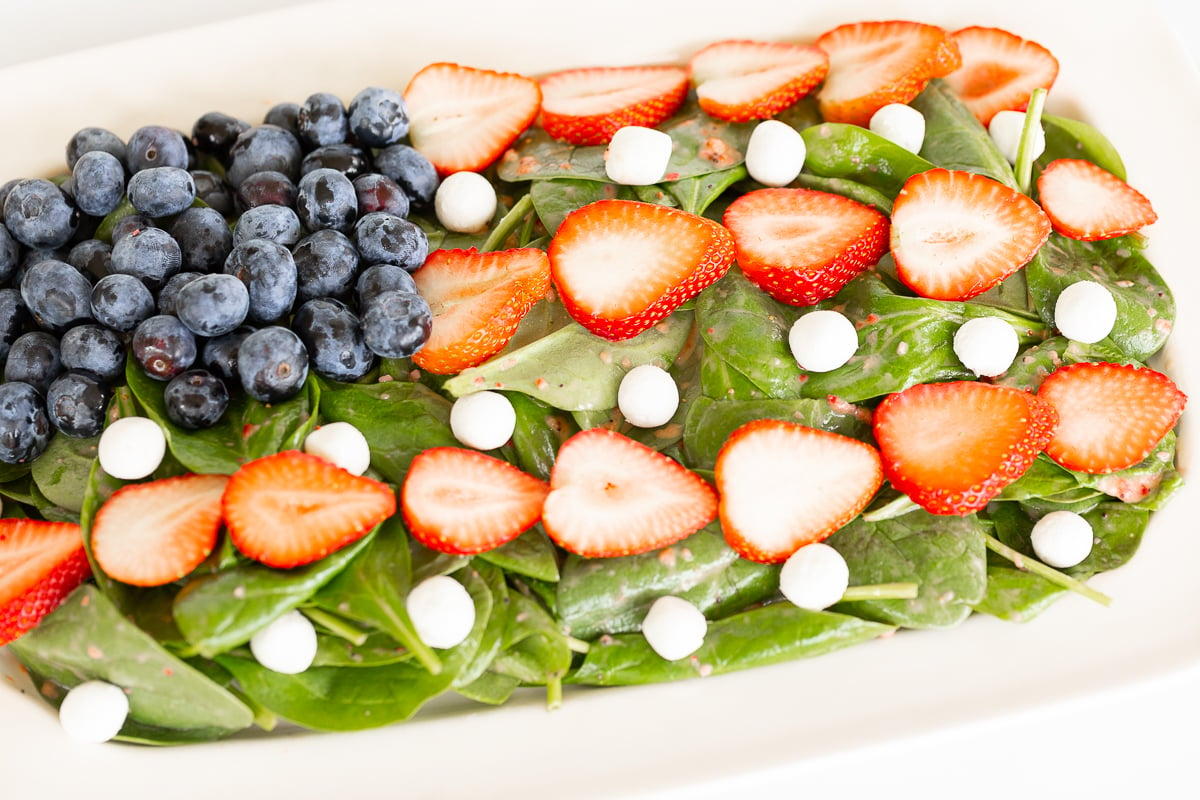 A fresh spinach salad topped with sliced strawberries, whole blueberries, and white cheese balls, served in a red dish.