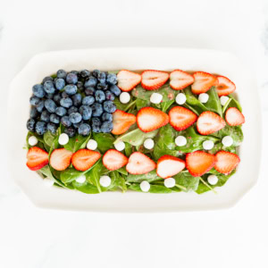 A rectangular platter with a red, white, and blue salad consisting of spinach topped with sliced strawberries, whole blueberries, and small white cheese pieces.